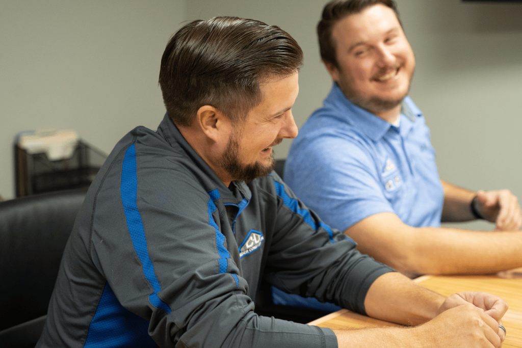 This image shows two men in a casual office environment, both wearing collared shirts with the logo of their organization on the chest. The man in the foreground, with neatly styled hair and a beard, is laughing and turning towards his colleague, who is slightly out of focus in the background. The background is indistinct, suggesting a typical office setting.