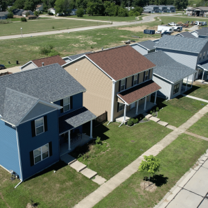 An aerial view of a suburban street featuring a mix of two-story homes with different siding colors including blue, tan, and gray. Each home has a neatly kept lawn, and the neighborhood appears spacious with wide streets and scattered green areas.