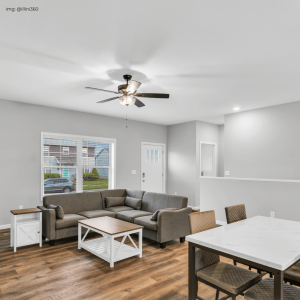 A bright, modern living space with a large sectional sofa and a white coffee table, set on rich hardwood flooring. The room features a ceiling fan, recessed lighting, and a dining area with a white table and patterned chairs. A large window offers a view of the neighborhood outside.