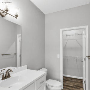 A clean, modern bathroom featuring a white vanity with a built-in sink, a bronze faucet, and a matching wall-mounted light fixture. The room has soft gray walls, a toilet to the side, and an open door leading to a small walk-in closet with built-in shelves and a wooden floor.