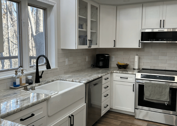 Bright, modern kitchen with white cabinets, marble countertops, a farmhouse sink, stainless steel appliances, and a large window overlooking a wooded view.