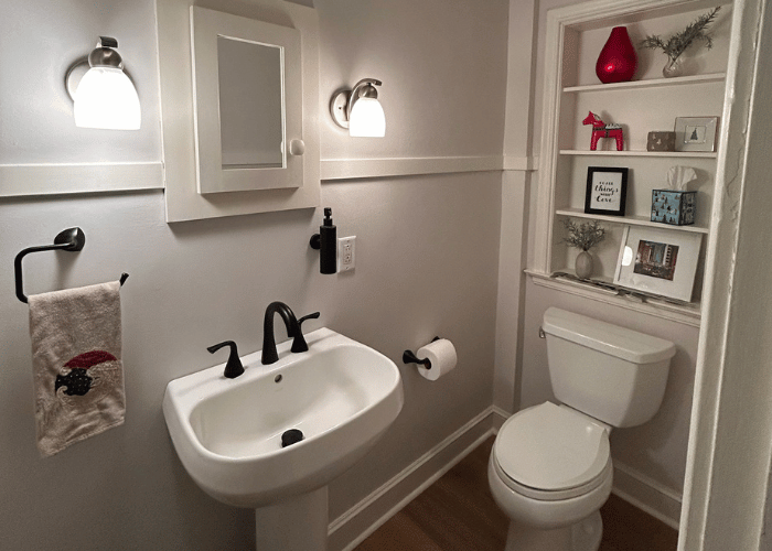 Modern small bathroom with a white pedestal sink, black faucet, wall-mounted mirror cabinet, and a toilet. Built-in shelving above the toilet holds decorative items, framed quotes, and plants. The space features soft white walls, warm wood flooring, and black hardware accents.